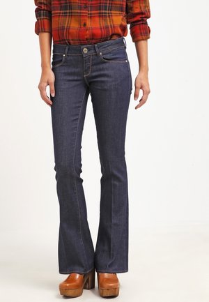 Donkerblauwe flared jeans met een gladde denimtextuur en voorzakken, gecombineerd met bruine platform schoenen. Bovenkleding is een rood-oranje geruite shirt.