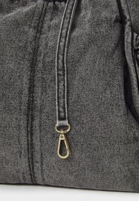 Nahaufnahme von grauem Denimstoff mit schwarzem Nähen und einem messingfarbenen Metallverschluss, der an einem passenden Riemen befestigt ist.