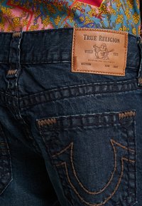 Mörkblå denimjeans har en läderlapp med "True Religion"-branding, sömmar på bakfickorna och kontrasterande bruna trådar.