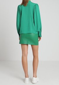Blouse verte à motifs avec des manches bouffantes, assortie à une mini jupe en cuir vert moulante. Le mannequin porte des baskets blanches.