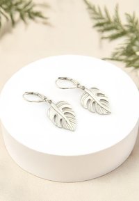 Smycken i form av silverörhängen med utstansade detaljer, hängande från vippklämmor, presenterade på en cirkulär vit display.