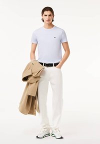 Camiseta azul clara, pantalones blancos y una chaqueta beige sobre el brazo. Conjunto complementado con zapatillas blancas con acentos verdes y negros.