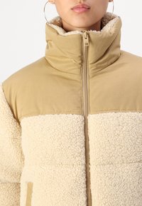 Beżowa i kremowa kurtka na zamek błyskawiczny, charakteryzująca się miękką, puszystą fakturą, wysokim kołnierzem oraz połączeniem gładkiej i teksturowanej tkaniny. Brak widocznych grafik lub ozdób.
