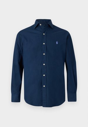 Jonge man staat frontaal, draagt een bruine pet, een navy blouse met knopen, donkere broek en meerkleurige bruine, groene en blauwe schoenen.