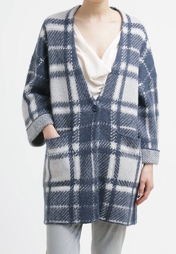 Femme portant un long manteau en laine à carreaux bleu et gris par-dessus un chemisier blanc drapé, une main dans la poche, et un pantalon gris clair.