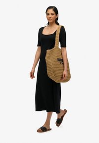 Sac en paille tressée avec un corps large et texturé, présentant un logo imprimé en bleu foncé. Le mannequin porte une robe noire ajustée et des sandales.