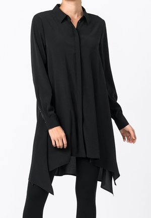 Langes schwarzes Hemdblusenkleid mit spitzem, asymmetrischem Saum, getragen über schwarzen Leggings, mit Kragen und langen Ärmeln mit geknöpften Manschetten.