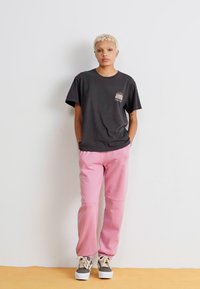 Black short-sleeve t-shirt with a graphic on the chest paired with light pink relaxed-fit pants and beige sneakers with white soles.