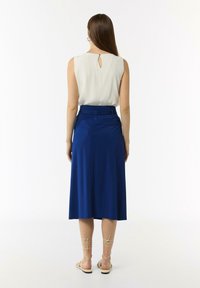 Ärmelloses cremefarbenes Top mit Rückenschlitzdetails, kombiniert mit einem mittellangen blauen Rock, der einen elastischen Bund und eine glatte Textur aufweist.