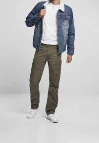 Hombre con zapatillas blancas, pantalones cargo oliva, camiseta blanca y chaqueta de denim azul con cuello de forro polar blanco, de pie frente a un fondo gris.