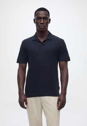 Junger Mann steht frontal, trägt ein schwarzes Polohemd mit kurzen Ärmeln und beige Hose vor einem schlichten weißen Hintergrund.