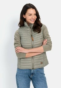 Jasnooliwkowa kamizelka puchowa z wysokim kołnierzem, zapięciem na zamek i ściągaczami na rękawach. Szczególnymi detalami są skórzana metka oraz pikowany wzór.