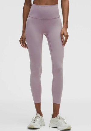 Person iført højtaljede støvede lilla leggings og hvide sports sko, stående mod en ensfarvet lys baggrund.