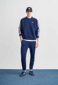 Sweatshirt azul-marinho com design de três riscas brancas, calças azul-marinho a condizer, sapatos azul-marinho e boné preto com logotipo branco. Apresenta um estilo clássico de roupa desportiva.