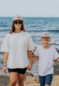 Dva posameznika na plaži, ki se držita za roke. Ženska nosi ohlapen bel srajc, črne kratke hlače in sivo kapo z oranžnim akcentom. Fant je oblečen v belo srajco in modre kratke hlače.