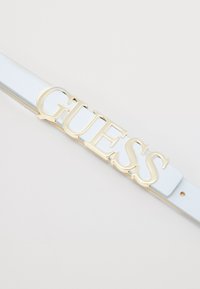Lyseblå faux skinnbelte med en gullfarget "GUESS" logo plassert foran og i midten. Glatt tekstur med standard spenne.