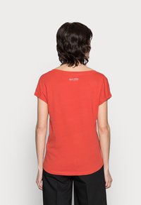 Rød kortærmet t-shirt med rund halsudskæring og lille hvidt logo på ryggen, lavet af blødt bomuldsstof. Glat tekstur.