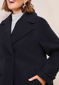 Femme souriante, portant de grandes boucles d'oreilles en argent, un col roulé côtelé gris et un manteau noir croisé avec sa main dans la poche.
