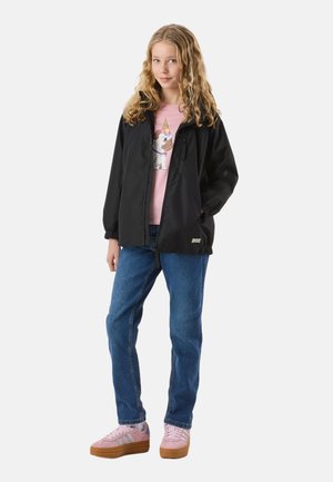 Ung pige med langt krøllet hår iført en sort jakke, lyserød T-shirt med hundemotiv, blå jeans og lyserøde sneakers, stående med den ene hånd i lommen.