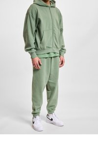 Groene ritssluiting hoodie met voorzakken en bijpassende sweatpants met cuffs, beide gemaakt van zachte stof; gedragen met witte sneakers met zwarte accenten.