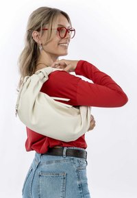 Kremowa torba na ramię o miękkiej teksturze i szczególe w postaci węzła na pasku. Nosi ją na czerwonej bluzce z długim rękawem oraz niebieskich dżinsach.