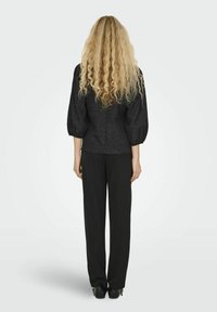 Blusa nera texturizzata con maniche a sbuffo abbinata a pantaloni neri dritti e scarpe nere. La vista posteriore mostra capelli lunghi e mossi.