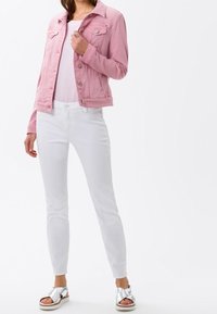 Nainen, joka on pukeutunut vaaleanpunaiseen denimtakkiin, valkoiseen t-paitaan, valkoisiin skinny-farkkuihin ja hopeisiin korkokenkiin, seisoo valkoista taustaa vasten.