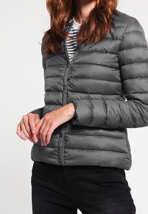 Personne portant une veste matelassée grise zippée sur une chemise rayée, les mains jointes devant et un pantalon noir visible.