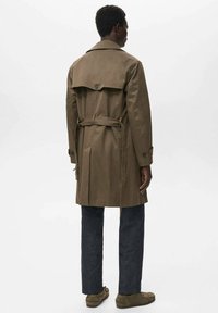 Een bruin trenchcoat met een ceintuur in de taille, een rugventilatieopening en knoopschouderepauletten. Draagt over donkerblauwe jeans en bruine schoenen.