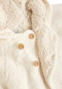 Ivory värvi fliiskate, millel on pehme ja udune tekstuur; omaduseks puidust nööbid ja mugav kapuuts. Silt on nähtav õmbluse lähedal.