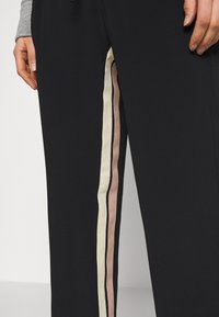 Svarta träningsbyxor med sidostripes i kräm och ljusrosa, med en slät yta och smal passform. Nära bild av en hand som vilar på tyget.