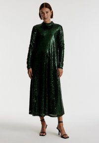 Groene pailletten maxi-jurk met lange mouwen en hoge hals; heeft een vloeiende silhouet en een gladde textuur, gecombineerd met zwarte strappy hakken.