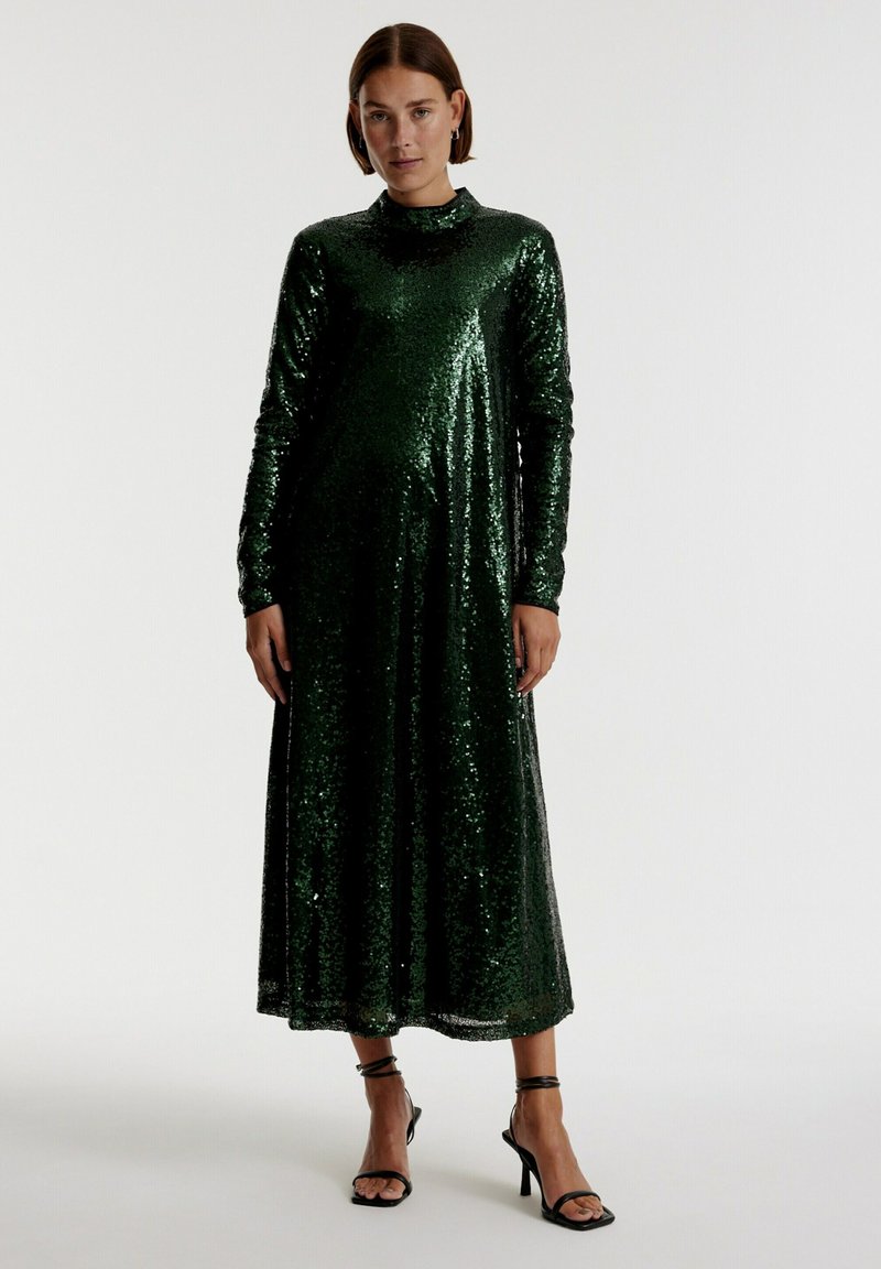 Groene pailletten maxi-jurk met lange mouwen en hoge hals; heeft een vloeiende silhouet en een gladde textuur, gecombineerd met zwarte strappy hakken.