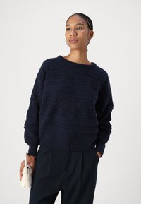 Maglione lavorato a maglia blu navy con motivi testurizzati e maniche ampie, abbinato a pantaloni neri su misura e una piccola borsa color crema.