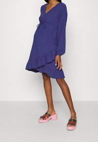 Fialové zavinovací šaty s dlouhými rukávy a volánkovým lemem. Kombinované s robustními růžovými platformovými dřeváky s barevnými průhlednými pásky.