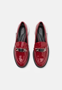 Punaiset lakatut loafersit, joissa on musta DKNY logo ja hopeinen ketjuyksityiskohta. Pyöreä varvas ja kumipohja. Sileä pinta, moderni muotoilu.