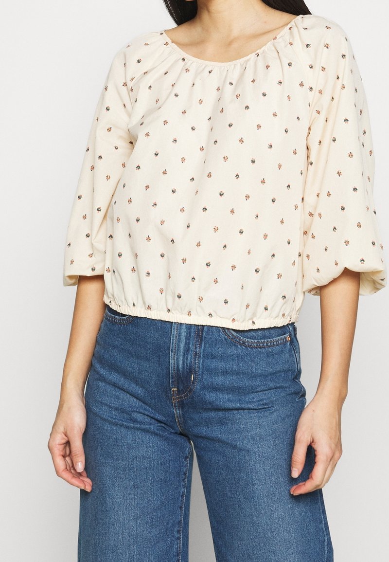 Femme portant un chemisier crème à petits motifs floraux et manches trois-quarts, associé à un jean bleu taille haute.