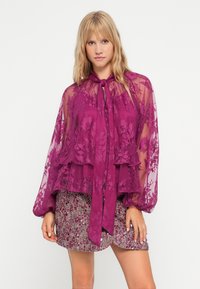 Bluză semi-transparentă magenta cu model din dantelă florală, mâneci tip balon și decolteu cu cravată; designul stratificat completează o fustă texturată, multicoloră.