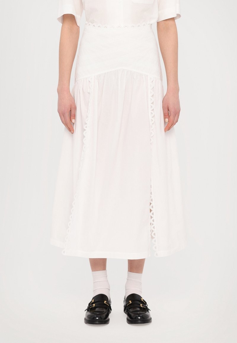 Jupe midi blanche avec bordure festonnée, taille froncée et tissu fluide ; portée avec des mocassins noirs et des chaussettes blanches montantes.