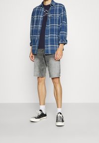 Mies, który ma na sobie niebieską koszulę w kratę na czarnym t-shircie z grafiką, szare szorty dżinsowe, białe skarpetki i czarne sneakersy na jednolitym tle.