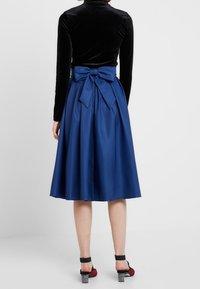 Sininen satiini midi-hame, jossa on leveä muoto, suuri takakujan yksityiskohtana ja sileä pinta. Yhdistettynä mustiin ja punaisiin korkokenkiin.