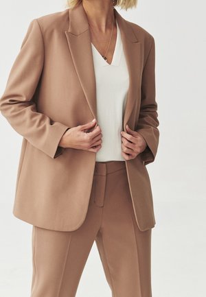 Persona que lleva un blazer beige y pantalones a juego con una camiseta blanca de cuello en V y un collar dorado, ajustando el frente del blazer.