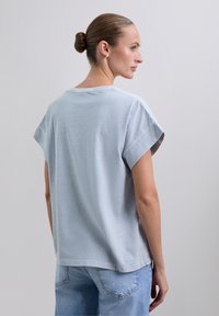 Frau mit einem Dutt, die ein hellgraues Kurzarmhemd und eine hellblaue Jeans trägt, leicht nach rechts gedreht vor einem einfarbigen Hintergrund.