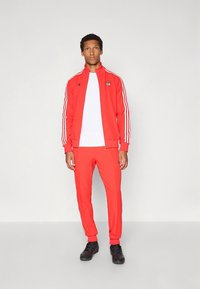 Röd atletisk träningsoverall i mjukt material med vita ränder på ärmarna och sidorna, med en logotyp på jackan.