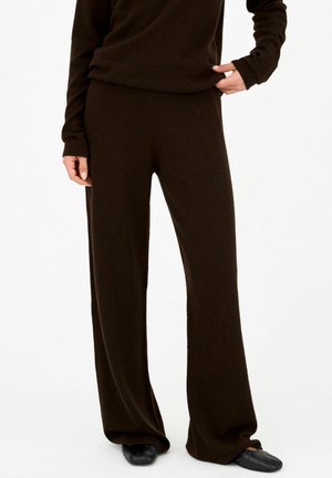 Personne portant un pantalon ample marron foncé et un haut à manches longues assorti, debout avec une main tenant l'ourlet du haut, chaussures noires visibles.
