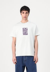 T-shirt en coton gris clair avec un col rond et des manches courtes, arborant un motif de fleur abstraite violette sur le devant.