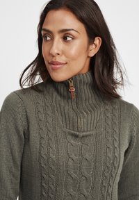 Dark green knitted sweater with a high collar, featuring cable knit patterns and a zipper at the neck. Textured, warm fabric.