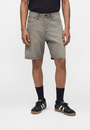 Muškarac obučen u sive traper kratke hlače, crne čarape i crne tenisice s bijelim prugama, stoji na jednostavnoj pozadini.