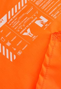 Leuchtend orangefarbener Stoff mit weißen gedruckten Notfallsignalen und Anweisungen. Merkmale sind Piktogramme für Hilfssignale und Gepäckprüfungen.