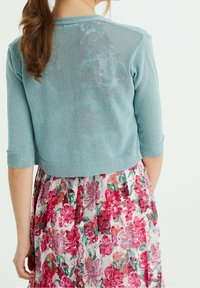 Světlomodrý krátký cardigan s tříčtvrtečními rukávy, vyrobený z třpytivé látky. Nositelný přes květinové šaty s růžovými a zelenými odstíny.